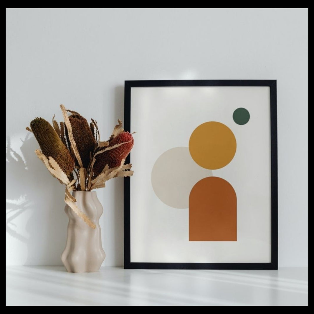 Framed abstract art print with geometric shapes next to a vase with dried plants on a white surface.